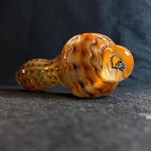 May include: A hand-blown glass pipe with a swirling pattern of orange, brown, and black. The pipe has a rounded bowl and a long, cylindrical stem. The mouthpiece is a clear, rounded shape with a black design.