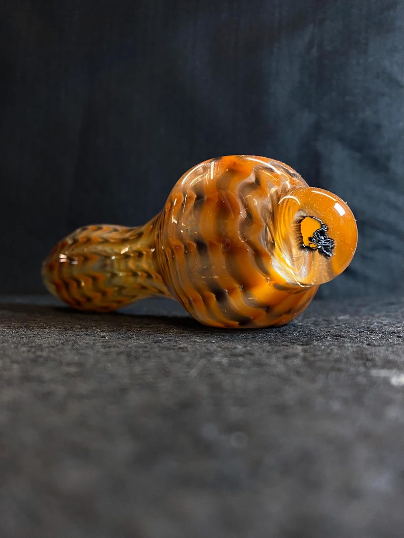 May include: A hand-blown glass pipe with a swirling pattern of orange, brown, and black. The pipe has a rounded bowl and a long, cylindrical stem. The mouthpiece is a clear, rounded shape with a black design.