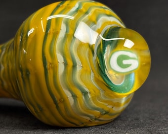 May include: A hand-blown glass pipe with a yellow, green, and white swirl design. The bowl features the letter "G" in green and white. The pipe is a unique, handcrafted piece.