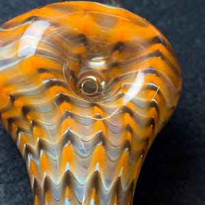 May include: Close-up of a hand-blown glass pipe with a vibrant orange, brown, and white swirl pattern. The pipe features a rounded bowl and a cylindrical stem. The glass has a glossy finish and a small hole.