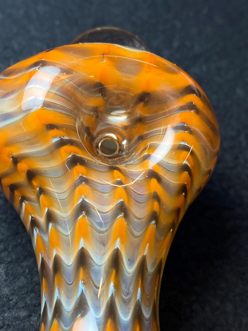 May include: Close-up of a hand-blown glass pipe with a vibrant orange, brown, and white swirl pattern. The pipe features a rounded bowl and a cylindrical stem. The glass has a glossy finish and a small hole.