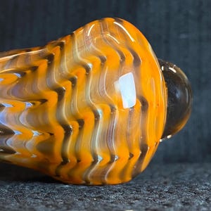 May include: Close-up of a glass pipe with a vibrant orange and black zigzag pattern. The pipe has a rounded bowl and a small, dark-colored mouthpiece. The glass appears to be translucent, with light reflecting off its surface.