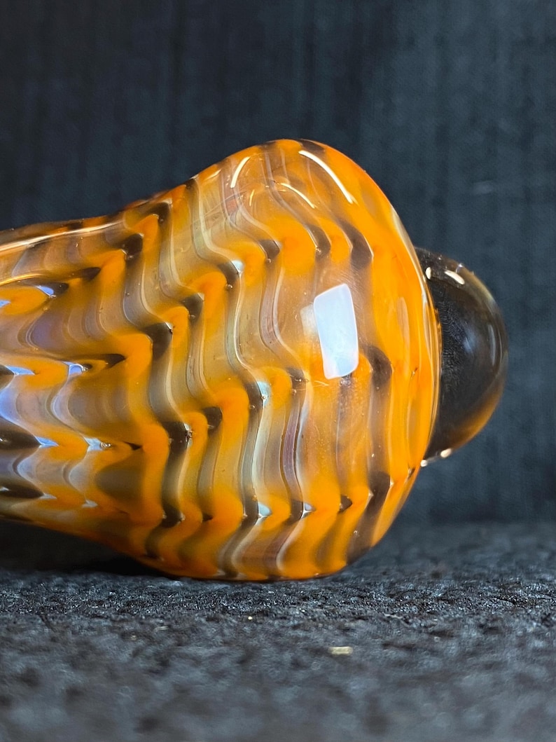 May include: Close-up of a glass pipe with a vibrant orange and black zigzag pattern. The pipe has a rounded bowl and a small, dark-colored mouthpiece. The glass appears to be translucent, with light reflecting off its surface.