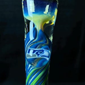 May include: A glass pipe with a blue, green, and yellow design. The pipe features a Seattle Seahawks logo in a blue and white bubble. The stem has a spiral pattern of blue and green stripes.