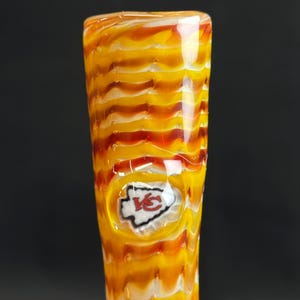 May include: A glass art piece with a yellow, orange, and red swirled design. The piece features the Kansas City Chiefs logo in the center. The glass has a textured, wavy pattern.