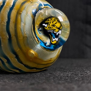 May include: A hand-blown glass pipe with a swirling pattern of gold and blue. The bowl features a detailed illustration of a jaguar's head with yellow, black, and white accents. The pipe is designed for smoking.