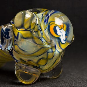 May include: A clear glass pipe with a swirling pattern of yellow, blue, and white. The pipe features a team logo with a white ram's head and an orange and blue background. The pipe has a rounded bowl and a curved stem.