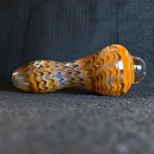May include: A glass pipe with an orange, brown, and blue patterned design. The pipe has a rounded bowl and a clear glass bubble on the end. The pipe is designed for smoking.
