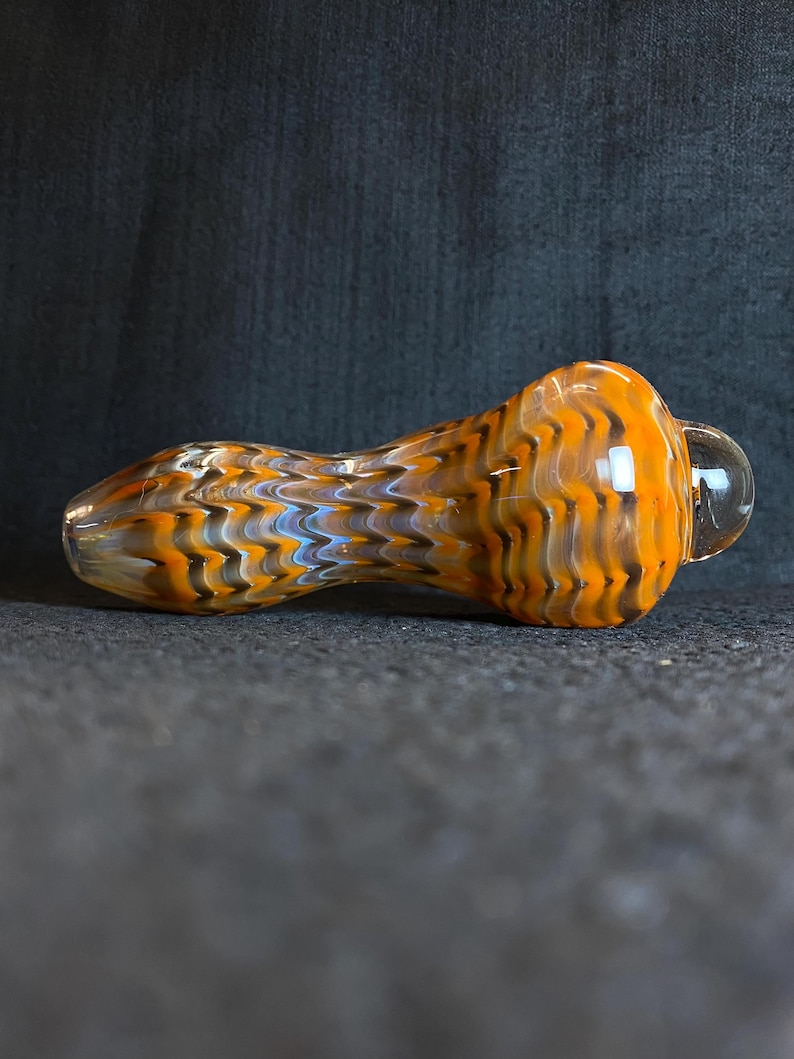 May include: A glass pipe with an orange, brown, and blue patterned design. The pipe has a rounded bowl and a clear glass bubble on the end. The pipe is designed for smoking.