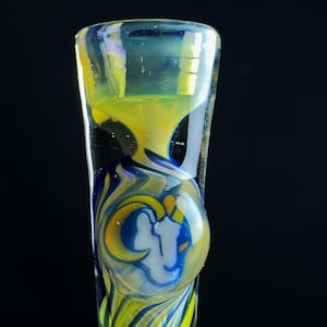 May include: A clear glass pipe with yellow, blue, and white swirling patterns. The design includes a round, raised section with a white and blue graphic. The top of the pipe has a flared opening.