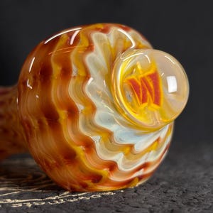 May include: A hand-blown glass pipe with a swirling pattern of orange, yellow, and white. The bowl features a clear, rounded design with a red and yellow interior. The pipe rests on a dark, textured surface.