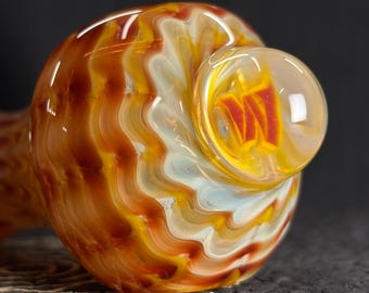 May include: A hand-blown glass pipe with a swirling pattern of orange, yellow, and white. The bowl features a clear, rounded design with a red and yellow interior. The pipe rests on a dark, textured surface.