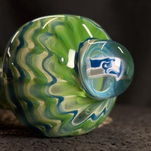 May include: A glass pipe with a round bowl and a smaller, spherical mouthpiece. The bowl features a swirling pattern of green, blue, and white. The mouthpiece has a blue and white design. The pipe is made of clear glass.