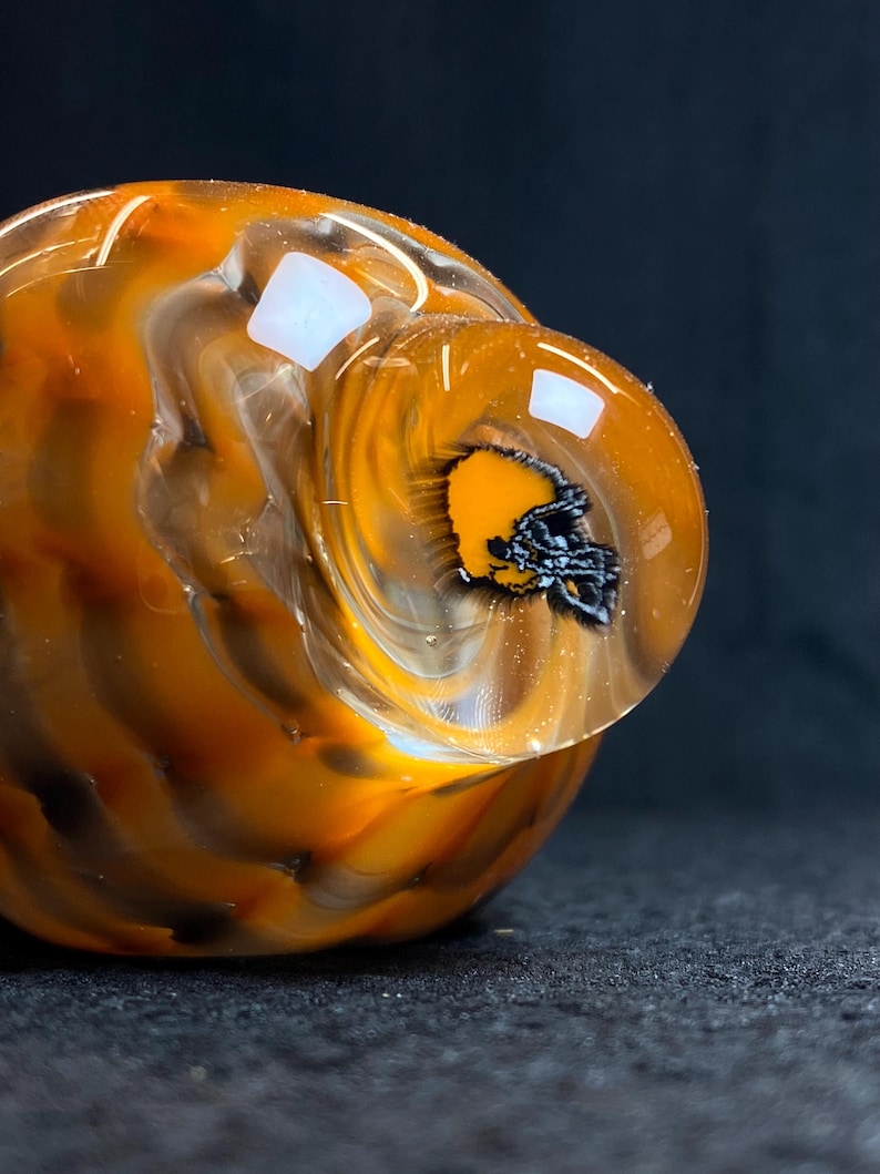 May include: A clear glass art piece with orange and black swirls. The piece has a rounded shape with a small, clear, circular extension. Inside the extension is a black and orange football helmet.