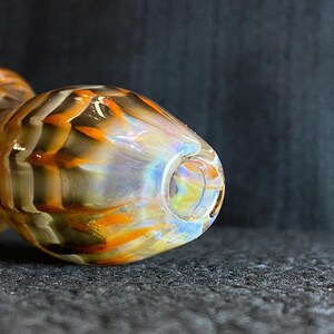 May include: A glass pipe with a rounded bowl and a stem. The pipe features a blend of colors, including orange, brown, and iridescent hues. The design has a textured appearance.
