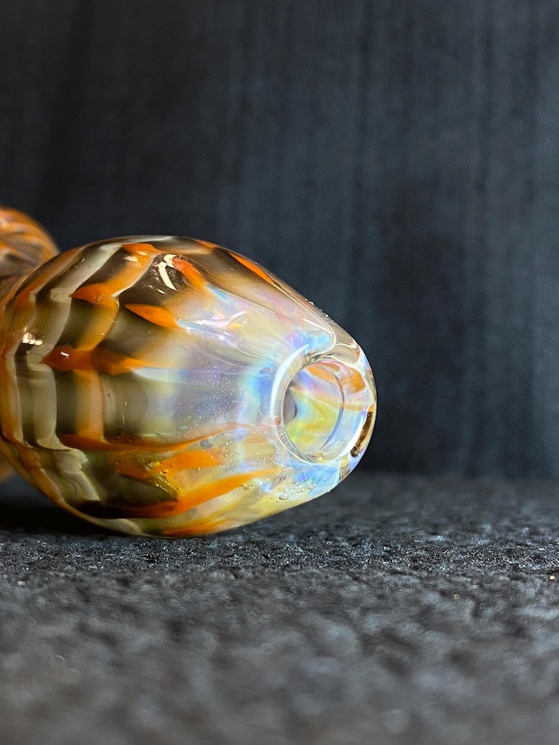 May include: A glass pipe with a rounded bowl and a stem. The pipe features a blend of colors, including orange, brown, and iridescent hues. The design has a textured appearance.