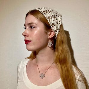 Puede incluir: Una diadema, pendientes y collar de ganchillo color crema con un colgante de flor. La joyería y la diadema son usados por una persona con cabello largo y claro y una camisa blanca.