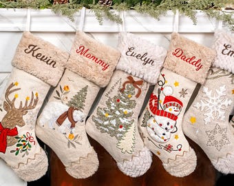 May include: Six personalized Christmas stockings hanging in a row, each featuring a beige fabric body, a plush faux fur cuff, and unique embroidered holiday designs including a reindeer, polar bear, tree, snowman, snowflake, and penguin.