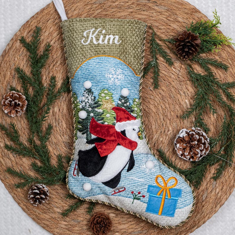 May include: A Christmas stocking featuring a textured olive green cuff with the embroidered name Kim. The body displays a winter scene with a penguin wearing a red Santa hat and scarf, surrounded by pine trees, snowflakes, and a blue gift box.