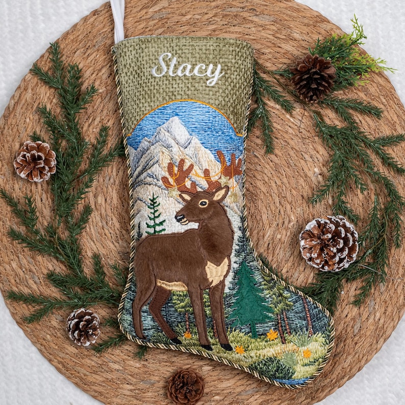 May include: A Christmas stocking made of tan burlap with a white embroidered name Stacy at the top. The stocking features a detailed embroidered scene of a brown deer standing in a forest with snow-capped mountains in the background.
