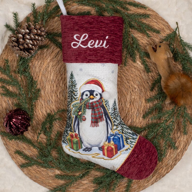 May include: A white Christmas stocking with a textured burgundy cuff embroidered with the name Levi in white script. The stocking features a central graphic of a penguin wearing a red hat and plaid scarf, surrounded by pine trees and gift boxes.