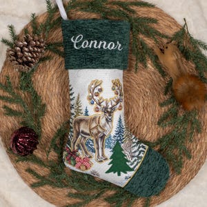 May include: A white Christmas stocking with a dark green cuff featuring the name Connor embroidered in white script. The stocking displays a reindeer with decorated antlers, surrounded by pine trees and gift boxes with red bows.
