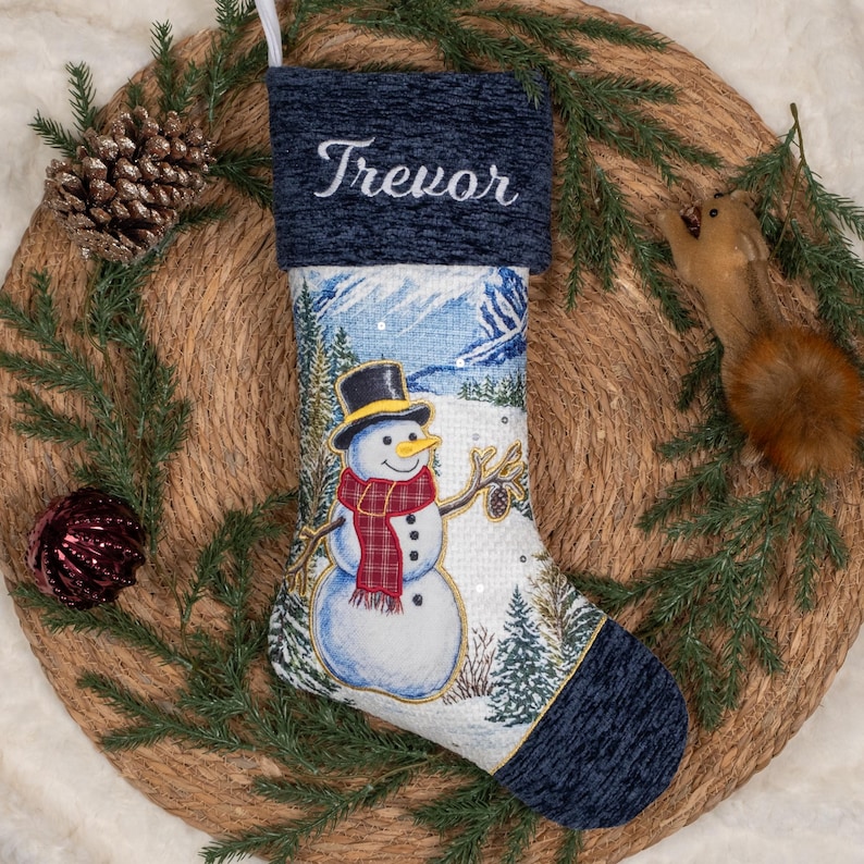 May include: A Christmas stocking featuring a dark blue textured cuff with the name Trevor embroidered in white script. The body displays a winter scene with a snowman wearing a black top hat and a red plaid scarf, surrounded by pine trees.