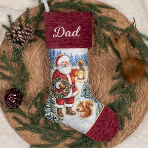 May include: A Christmas stocking featuring a deep red textured cuff with the embroidered word Dad. The body displays a printed scene of Santa Claus holding a lantern and a wreath in a snowy forest, accompanied by a squirrel near the toe.