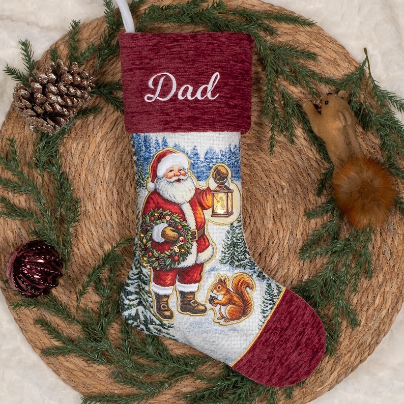 May include: A Christmas stocking featuring a deep red textured cuff with the embroidered word Dad. The body displays a printed scene of Santa Claus holding a lantern and a wreath in a snowy forest, accompanied by a squirrel near the toe.