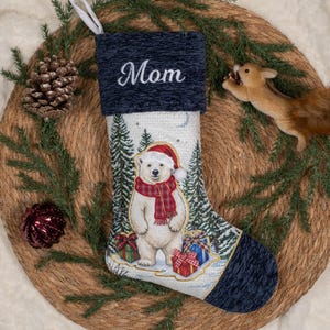 May include: A Christmas stocking with a dark blue textured cuff embroidered with the word Mom in white script. The body features a white polar bear wearing a red Santa hat and a red plaid scarf, surrounded by pine trees and colourful gift boxes.