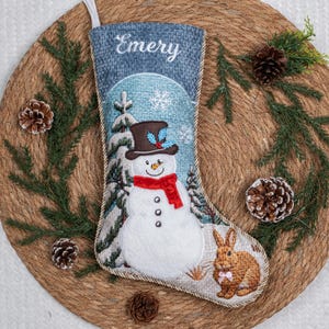 May include: A Christmas stocking featuring a blue textured top with the name Emery embroidered in white. The center displays an embroidered snowman wearing a brown top hat and red scarf, surrounded by snowy trees and a brown rabbit with a pink bow.