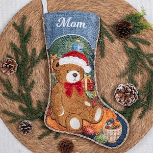 May include: A Christmas stocking featuring a brown teddy bear wearing a red Santa hat and a red bow tie. The top cuff is blue with the word Mom embroidered in white. The design includes a gift box and a bowl of ornaments on a textured blue background.