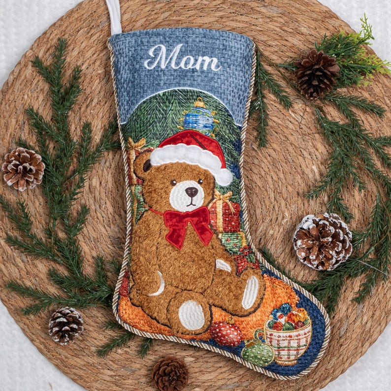 May include: A Christmas stocking featuring a brown teddy bear wearing a red Santa hat and a red bow tie. The top cuff is blue with the word Mom embroidered in white. The design includes a gift box and a bowl of ornaments on a textured blue background.