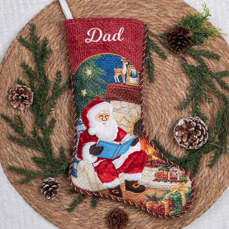 May include: A red Christmas stocking featuring the embroidered word Dad at the top. The design depicts Santa Claus sitting in a chair reading a blue book by a fireplace, with a small deer, a toy train, and wrapped gifts visible in the scene.