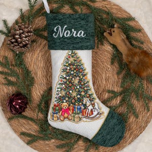 May include: A white Christmas stocking with a dark green textured cuff featuring the embroidered name Nora in white script. The stocking displays a colourful Christmas tree graphic with presents, a teddy bear, and a rocking horse toy.