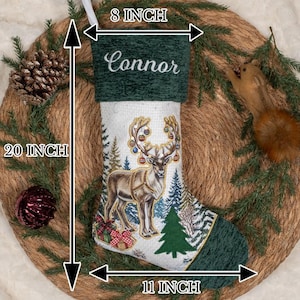 May include: A Christmas stocking measuring 50.8 cm in height, 20.3 cm at the top, and 27.9 cm at the base. It features a dark green cuff with the name Connor embroidered in white, and a white body with a reindeer, trees, and gifts.