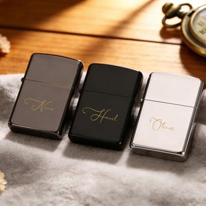 May include: Three personalized lighters in gunmetal, black, and silver. Each lighter has a name in elegant gold script: "Nina", "Hazel", and "Oliver". The lighters are displayed on a soft, gray surface with dried flowers and a pocket watch in the background.