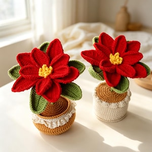 May include: Two crocheted poinsettia plants in pots. Each plant features vibrant red petals, a yellow center, and green leaves. The pots are crocheted in brown and cream colors, with one pot featuring a decorative white lace trim around the top edge.