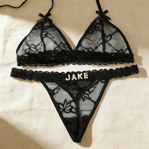 Op de afbeelding: Zwarte kanten lingerie set met een driehoekige bralette en een string. De bralette heeft delicate strikaccenten en de string heeft de naam "JAKE" in sprankelende letters. De set is gemaakt van doorschijnend, bloemmotief kant.
