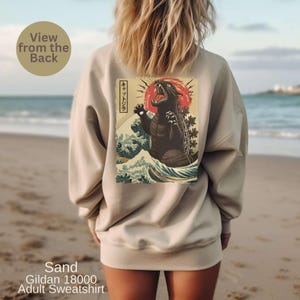 May include: Sand-colored adult sweatshirt featuring a Godzilla graphic in the style of a Japanese woodblock print. The design includes a red sun, waves, and Japanese text. The sweatshirt is a Gildan 18000.