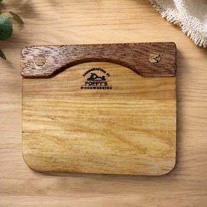 May include: A wooden dough scraper with a light wood base and a darker wood handle. The handle is curved and attached with two small pegs. The text "Handcrafted by Poppy's Woodworking" is printed on the base.