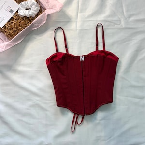 Op de afbeelding: Een bordeauxrode corset top met verstelbare bandjes en een sluiting aan de voorkant. De top heeft een satijnen rand en een strik aan de onderkant. Een witte scrunchie en een klein doosje met een kaart zijn ook zichtbaar.