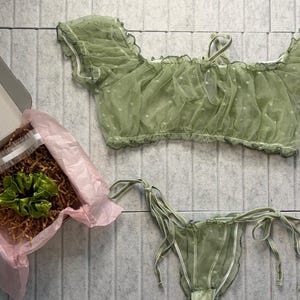 May include: A light green lingerie set, comprising a ruffled off-the-shoulder top and matching bottoms with tie details. The set is made of a sheer fabric with white polka dots. A gift box with a green plant is also visible.