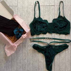 May include: An emerald green lingerie set, including a bra and thong, with a gift box containing black clothing and a teal scrunchie. The thong has the word "NAME" on the waistband. The set is displayed on a light grey tiled surface.