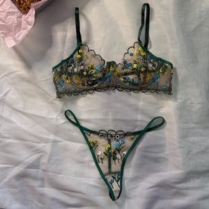May include: A lingerie set featuring a floral embroidered bra and thong with green trim. The bra and thong have a sheer, beige base with blue and yellow floral embroidery. A gift box with a scrunchie and a card that says "Thank You" is also included.