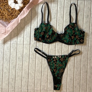 May include: Black lingerie set with green floral embroidery. The set includes a bra and a thong. The thong has the word "NAME" embroidered on it. The set is presented in a gift box with a scrunchie and a "Thank You" card.