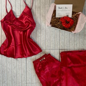 May include: A red satin pajama set with a camisole top and matching pants. The set includes a gift box with a scrunchie and a card that says "Thank You". The pajama pants have the word "NAME" embroidered on the waistband.