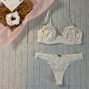 May include: A white floral print bra and matching thong underwear set with lace trim. The set is displayed on a gray tiled surface. A gift box with a scrunchie and a "Thank You" card is also visible.