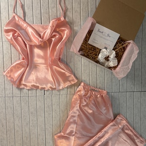 Op de afbeelding: Een lichtroze satijnen pyjamaset, bestaande uit een camisole top met spaghettibandjes en een broek. Een geschenkdoos met een scrunchie en een kaart met de tekst "Thank You" is ook inbegrepen.