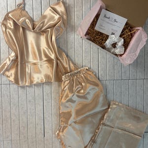 May include: A champagne-colored satin pajama set, including a camisole top with spaghetti straps and matching pants. A gift box with a "Thank You" card and a white scrunchie is also included. The set is displayed on a light gray tiled surface.